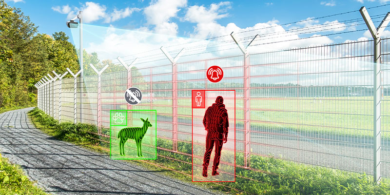 Perimeter fence with a surveillance camera and a dark human figure and a dark deer standing in front of it, both being detected by a surveillance camera